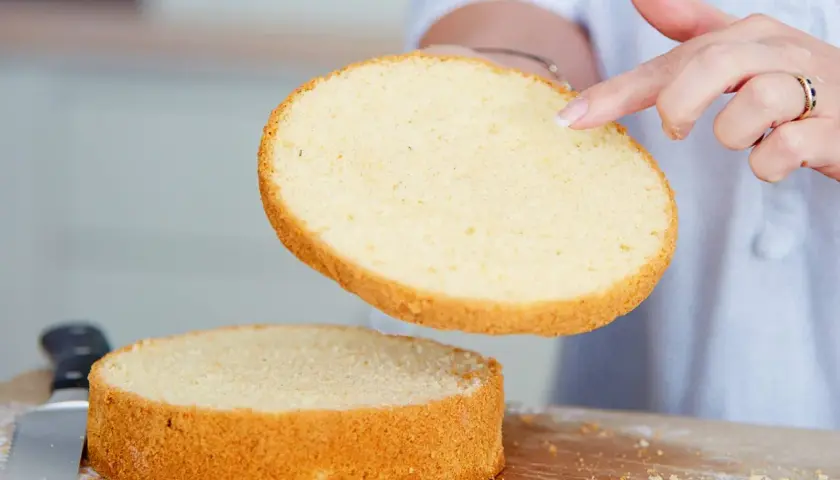 Perfekter tortenboden rezept: Eine Hand hebt eine goldbraune Kuchenschicht über eine zweite, die auf einem Holzbrett liegt.