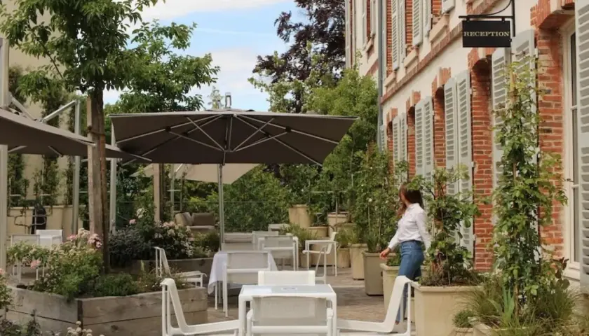 Terrasse verdoyante du 25bis by Leclerc Briant, avec tables, chaises et parasols. Une femme se dirige vers la réception.