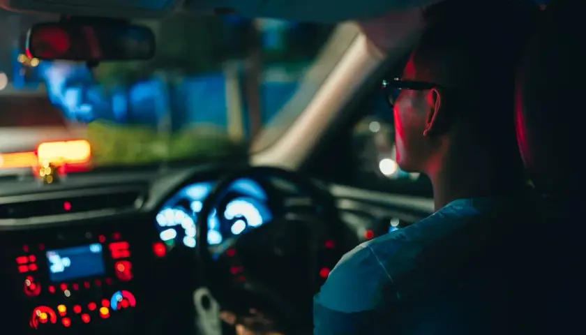 Un hombre con gafas conduce de noche, con el tablero iluminado en azul y rojo.