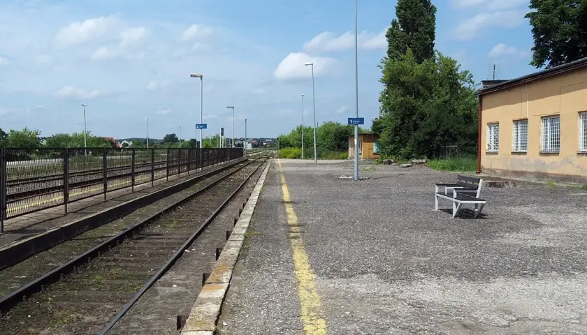 Pusta stacja kolejowa Opoczno Kielce. Torowisko, peron z ławką i żółtą linią, budynek dworca.