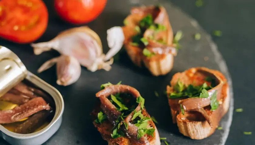Otwarta puszka anchois, bruschetty z anchois i natką pietruszki, pomidory i czosnek na ciemnym tle.