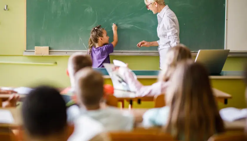 Eine Lehrerin steht vor einer Tafel, während ein Kind schreibt. Es ist schön, Lehrer zu sein und Wissen zu vermitteln.