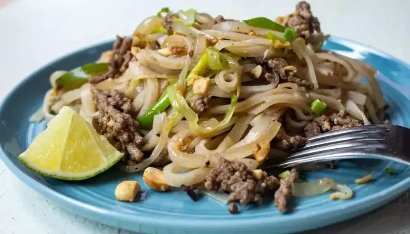 Pyszny pad thai przepis na talerzu: makaron ryżowy z mielonym mięsem, cebulą, orzeszkami ziemnymi i limonką.