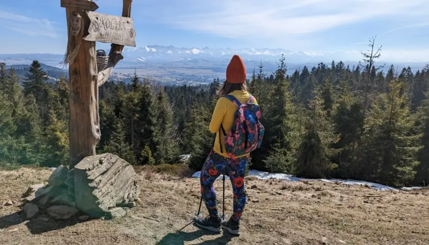 Widok na Tatry z drewnianego słupa z napisem "Koliba na Łapsowej Polanie". Turystka w kolorowych legginsach podziwia panoramę.