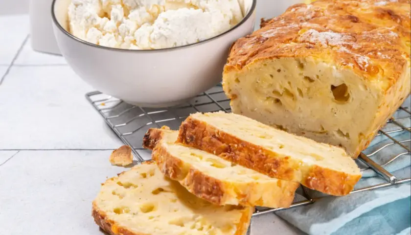 Chleb z twarogu, świeżo upieczony, z chrupiącą skórką i puszystym wnętrzem. Obok miska pełna białego twarogu, składnika tego pysznego wypieku.