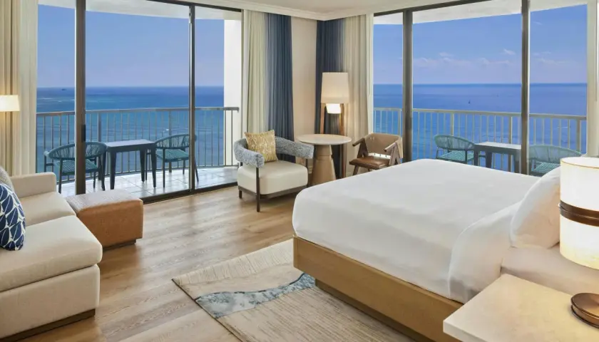 Pokój hotelowy z widokiem na ocean, idealny na wakacje hawaje all inclusive. Wygodne łóżko, sofa i balkon z stolikiem.