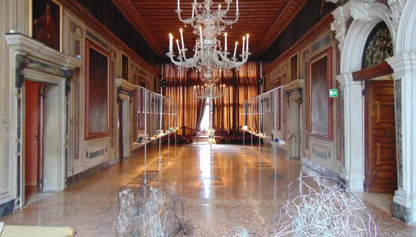 Intérieur du Palazzo Mocenigo, un long couloir orné de tableaux, avec un lustre imposant et des sculptures modernes au sol.