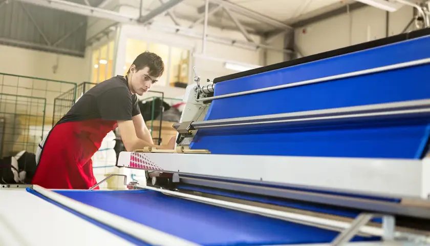 Un homme en tablier rouge travaille sur une machine textile bleue.