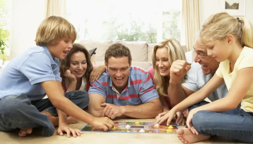 Familie spielt ein kooperatives Brettspiel, lachend und konzentriert auf dem Boden liegend.