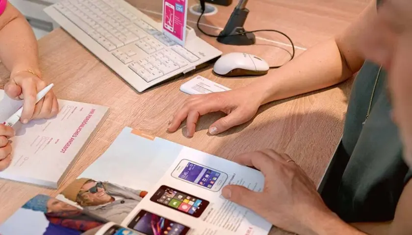 Klienci przeglądają ofertę smartfonów T-Mobile. Na biurku klawiatura, myszka i dokumenty.