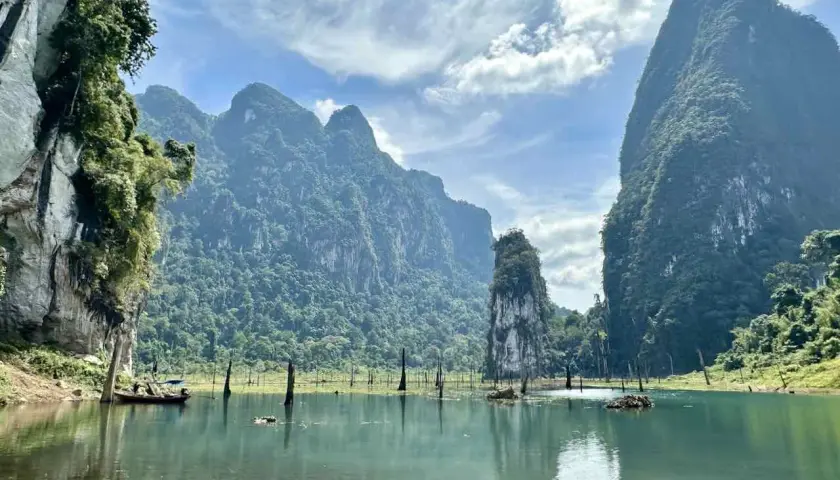 Khao Sok Nationalpark: Majestätische Kalksteinfelsen ragen aus einem ruhigen See empor, umgeben von üppigem Dschungel. Ein Boot gleitet über das Wasser.