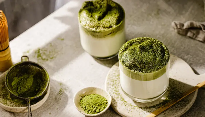 Zwei Gläser cremiger Matcha-Latte, bestreut mit feinem Pulver. Die Zubereitung mit Chasen und Sieb deutet auf die belebende matcha wirkung hin.