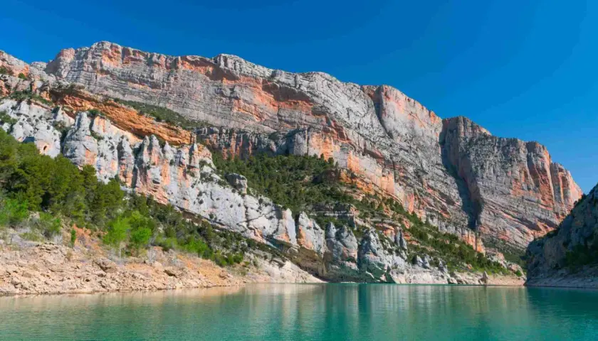 Majestuosas paredes rocosas de la sierra del Montsec se reflejan en las aguas turquesas de un embalse.