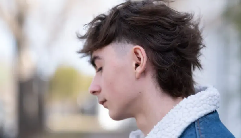Junger Mann mit modernen Mullets, der nachdenklich zur Seite blickt. Sein Haar ist dunkelbraun und zerzaust, die Seiten sind kurz geschoren. Er trägt eine Jeansjacke mit Teddyfellkragen.