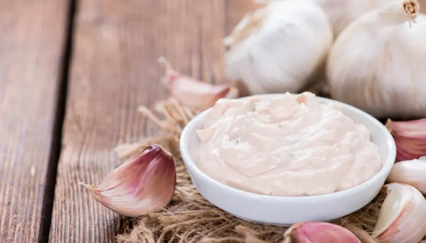Kremowy sos aioli w miseczce, otoczony świeżymi ząbkami czosnku i całymi główkami na drewnianym tle.