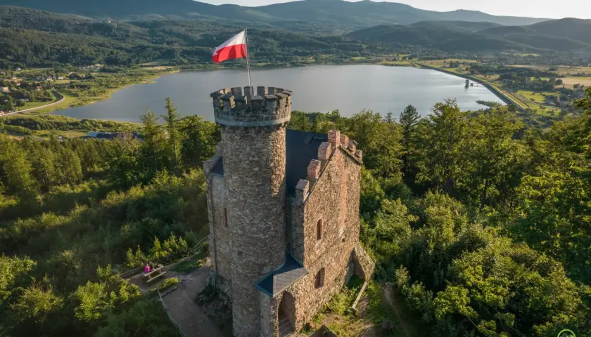 Kamienny zamek henryka z polską flagą na wieży, otoczony zielenią, z jeziorem i górami w tle.