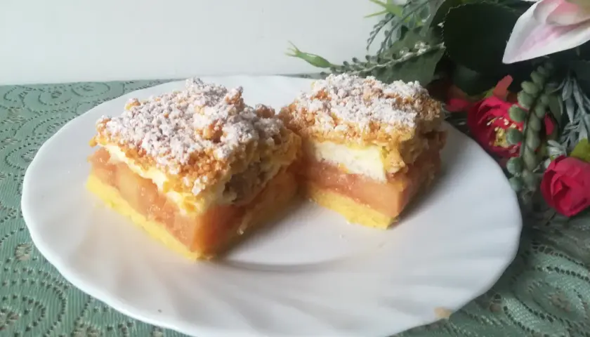 Dwa kawałki szarlotki z bezą i kruszonką, posypane cukrem pudrem, na białym talerzu.