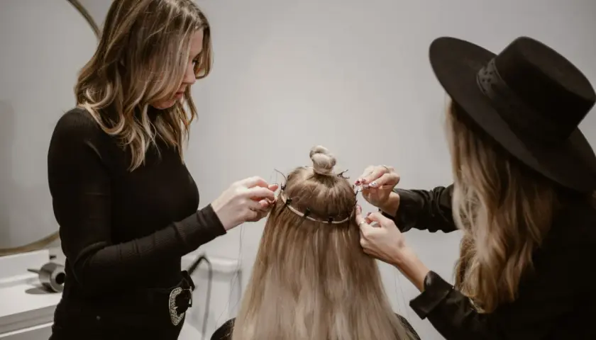 Två personer hjälper till att **slinga håret hemma** med extensions. En med svart polotröja och en med svart hatt arbetar på det långa blonda håret.