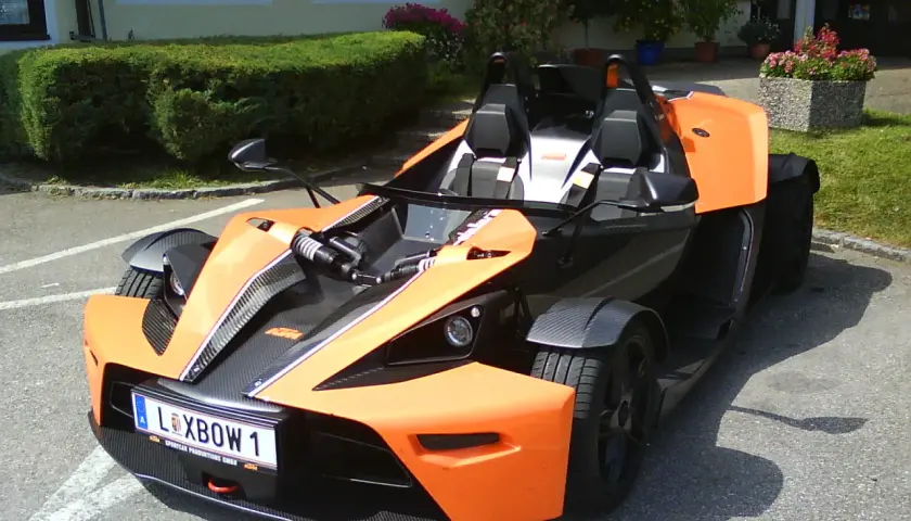 Pomarańczowy KTM X-Bow z czarnymi akcentami i tablicą rejestracyjną "XBOW 1" stoi na parkingu przed budynkiem z zielonym żywopłotem.