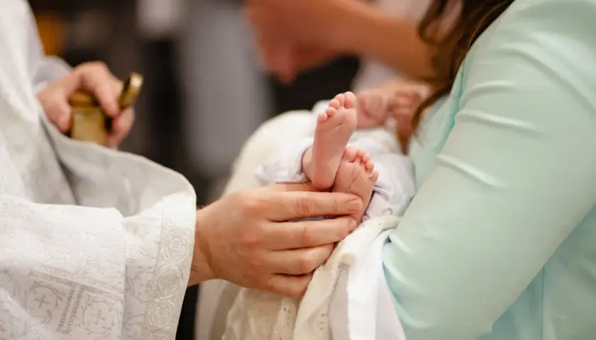 Chrzest dziecka, gdzie dziadkowie mogą być chrzestnymi. Dłoń trzyma stópki niemowlęcia, obok kapłan z naczyniem.