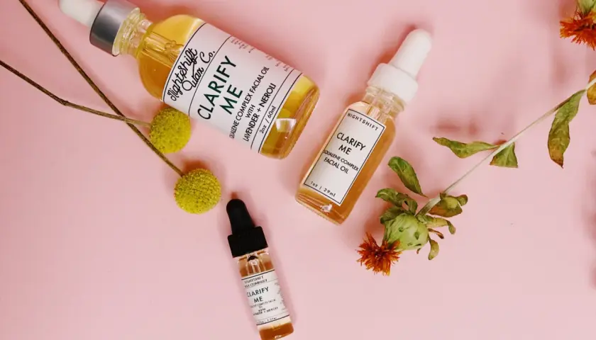 Tres frascos de aceite facial "Clarify Me" con escualeno, lavanda y neroli, rodeados de flores secas sobre fondo rosa.