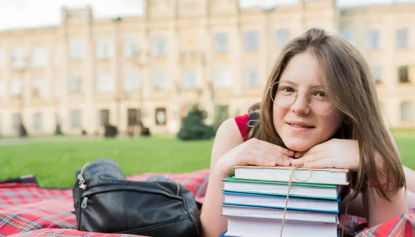Dziewczyna z okularami leży na kocu z książkami. Posiadanie certyfikatu C1 angielski otwiera drzwi do kariery i studiów.