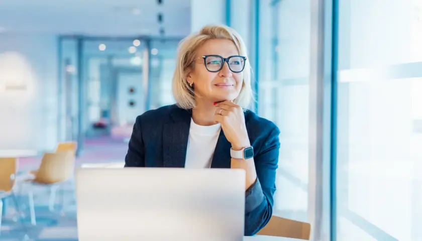 Donna bionda con occhiali, seduta al computer, pensa al suo futuro. Un nuovo capitolo, cambiare lavoro a 50 anni, è una possibilità concreta.