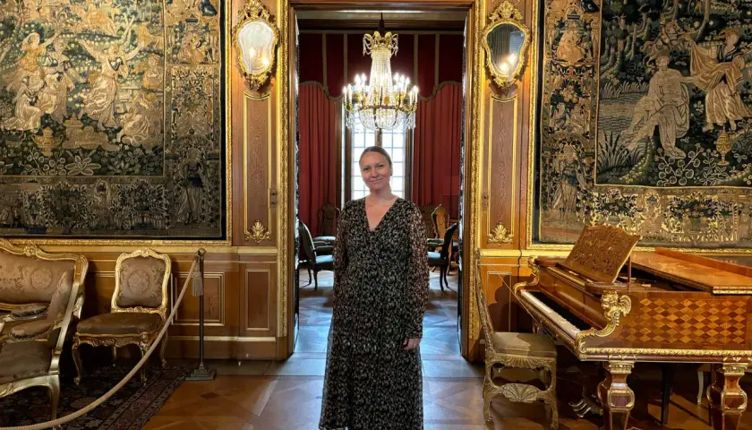 En kvinna står i en sal på Hallwylska museet Stockholm, omgiven av antika gobelänger, en flygel och utsökt inredning.