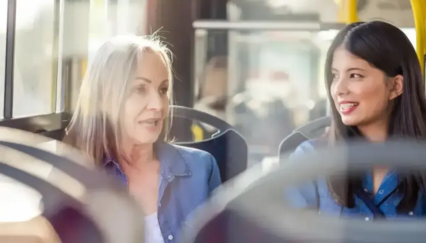 Dwie kobiety, starsza i młodsza, siedzą obok siebie w autobusie. Starsza, z siwymi włosami, opowiada coś młodszej, która słucha z uśmiechem. To miła rozmowa z nieznajomym.