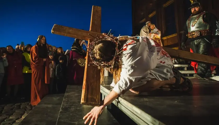 Scena kiedy jest Droga Krzyżowa: Jezus w cierniowej koronie, z krwią na szacie, upada pod ciężarem krzyża, podczas gdy tłum obserwuje.