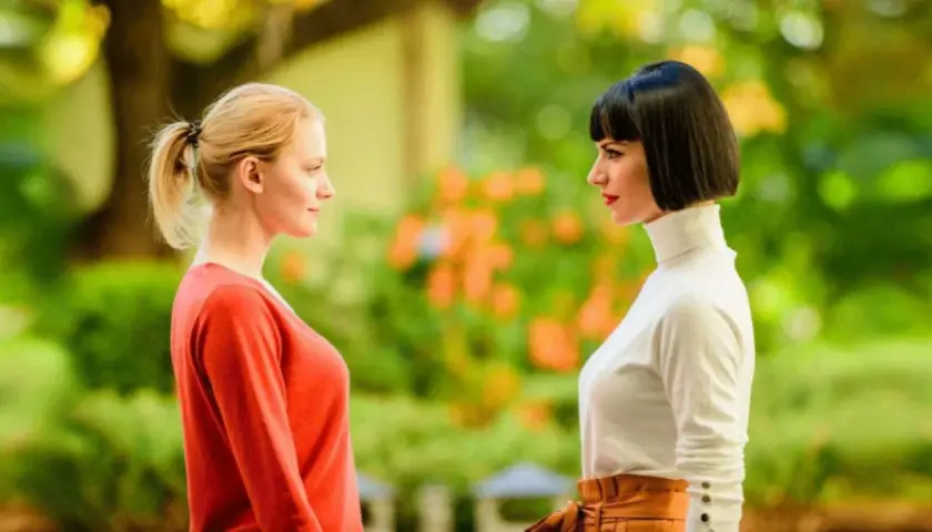 Due donne si guardano intensamente, una con i capelli biondi raccolti, l'altra con un caschetto scuro. Si percepisce un'atmosfera di invidia e gelosia tra loro.