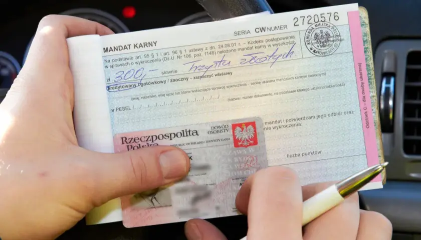 Ręka trzyma mandat karny na 300 zł za parkowanie na zakazie, obok dowód osobisty.