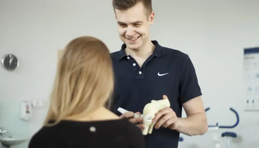 Uśmiechnięty technik fizjoterapii pokazuje pacjentce model stawu kolanowego, wyjaśniając szczegóły.