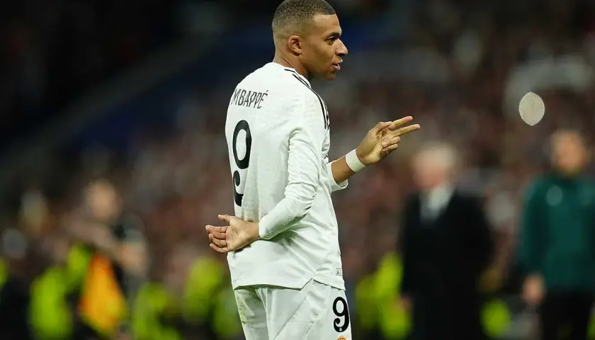 Mbappe, w białej koszulce z numerem 9, gestykuluje na boisku. Jego wzrost robi wrażenie, a kibice śledzą każdy jego ruch.