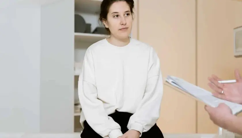 Kobieta w białej bluzie siedzi przed lekarzem trzymającym dokumenty.
