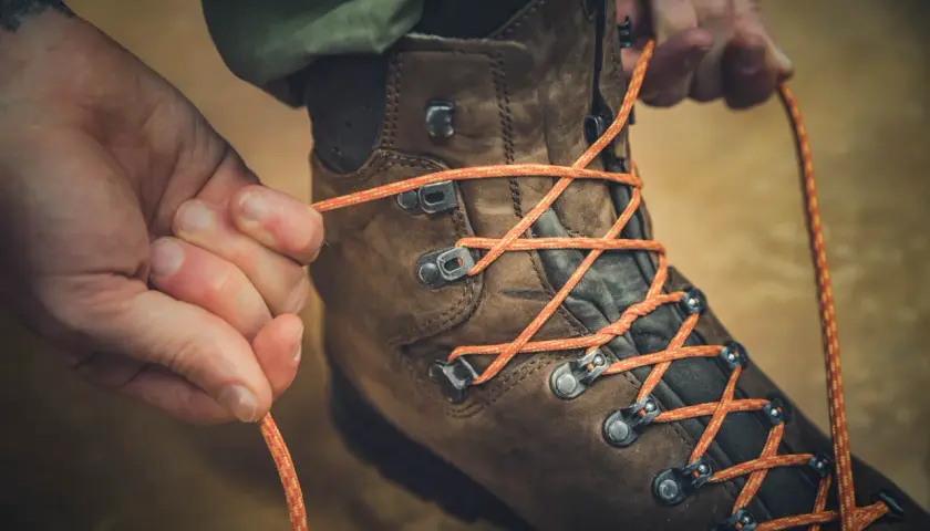 Close-up of hands tying orange laces through metal hooks on a brown hiking boot, demonstrating how to tie boots with hooks for a secure fit.