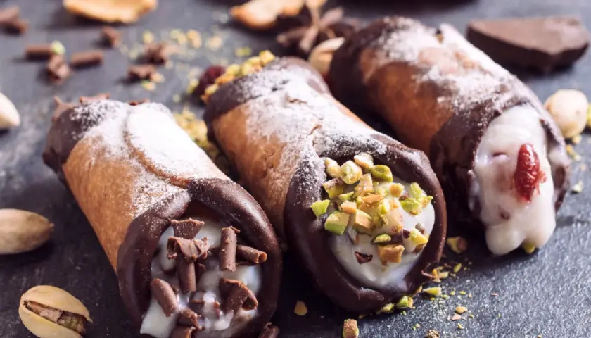 Trzy pyszne cannoli z kremem, posypane cukrem pudrem, pistacjami i czekoladą. Idealne na deser, gdy szukasz inspiracji na cannoli przepis.