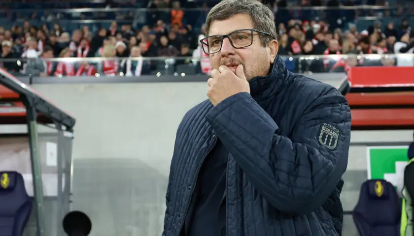 Włoski trener w okularach, zamyślony, w granatowej kurtce z naszywką "ITALIA", stoi na tle trybun stadionu.