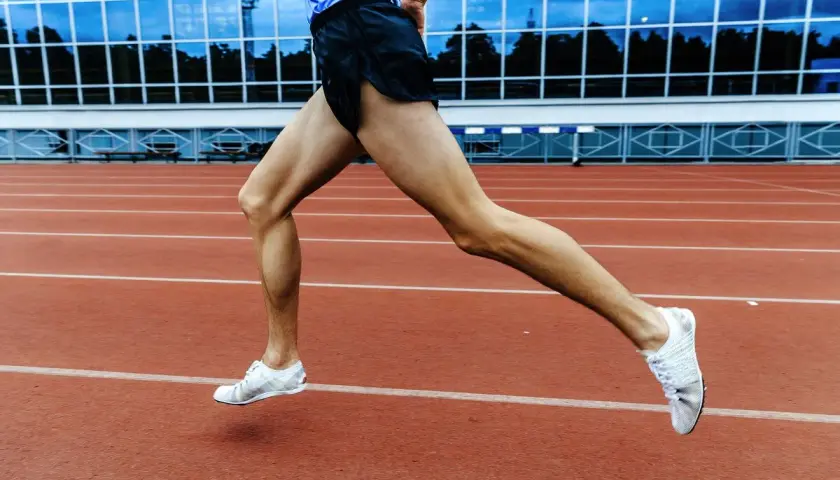 Sylwetka biegacza w ruchu na bieżni stadionu. Mięśnie nóg naprężone, białe buty sportowe unoszą się nad czerwonym torem.