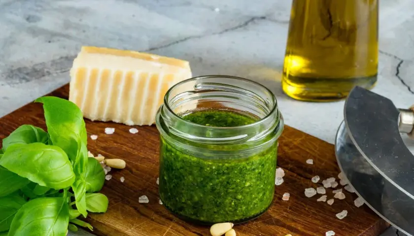 Słoiczek domowego pesto z bazylii, idealny sposób, jak zrobić pesto z bazylii na zimę. Obok świeża bazylia, orzeszki piniowe, parmezan i oliwa.