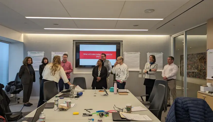 A team coach leads a meeting in a conference room, with a screen displaying "What will make today successful for you?".