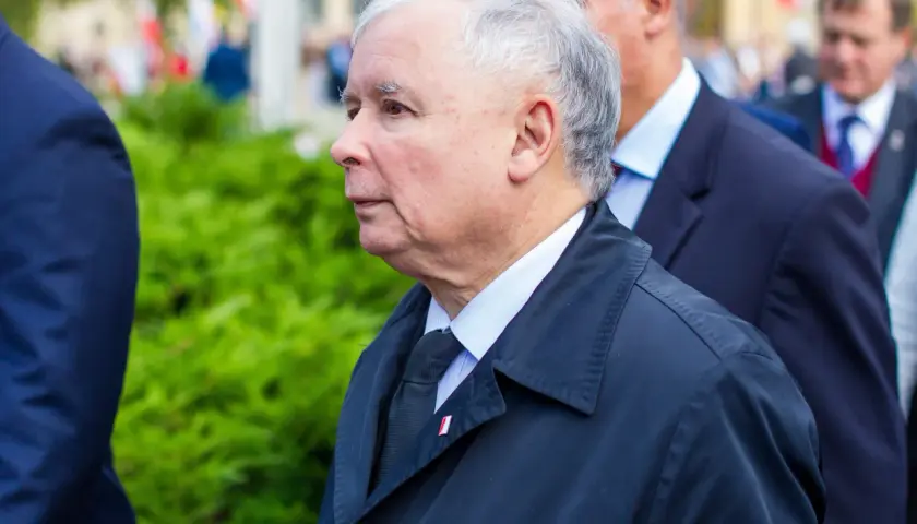 Jarosław Kaczyński, lider partii, w czarnym płaszczu i z przypinką z polską flagą, podczas oficjalnych uroczystości.