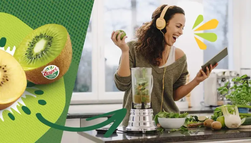 Glad kvinna med hörlurar gör smoothie med kiwi nyttigt. Två halverade kiwifrukter syns till vänster.