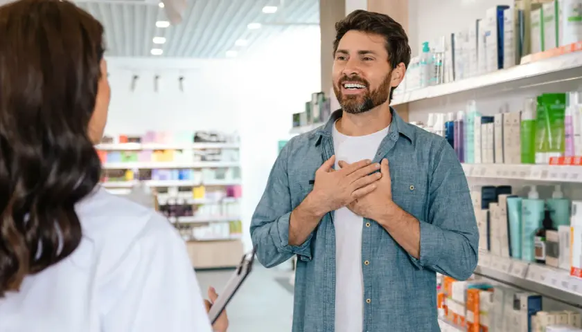 Mężczyzna z dłońmi na piersi rozmawia z farmaceutką, zastanawiając się, ile zarabia farmaceuta.