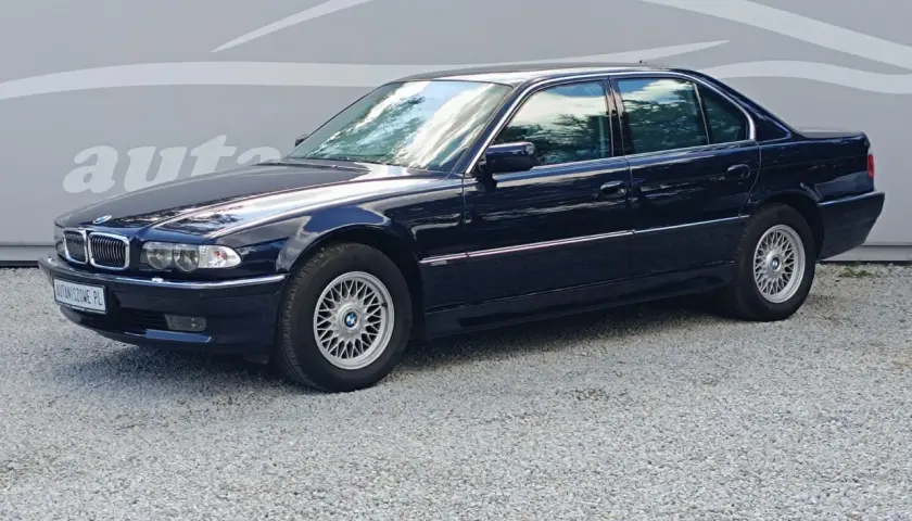 Ciemnoniebieskie BMW 7 E38 z lat 90. stoi na żwirowym podjeździe, z odbiciem chmur w szybach.