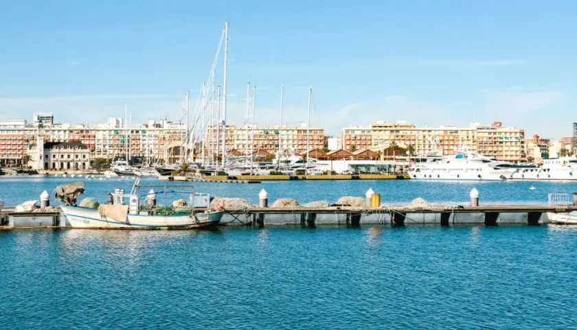 Barcos de pesca y yates de lujo atracados en la marina de Valencia, con edificios coloridos de fondo bajo un cielo azul.