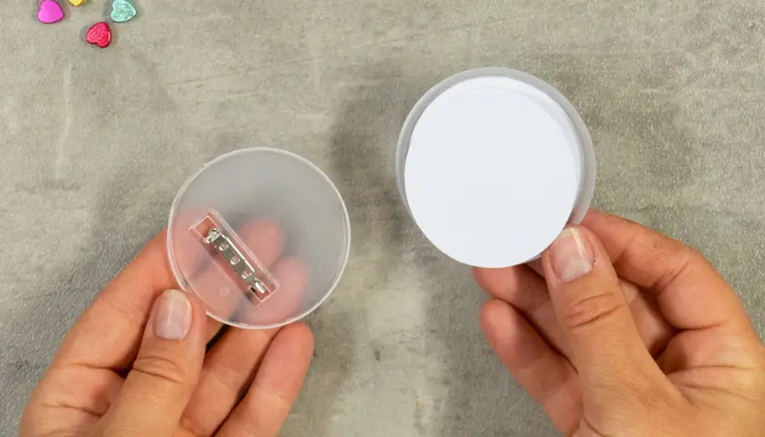 Zwei Hände halten Teile für das **button basteln**: eine mit einer transparenten Rückseite und Nadel, die andere mit einem weißen Kreis.