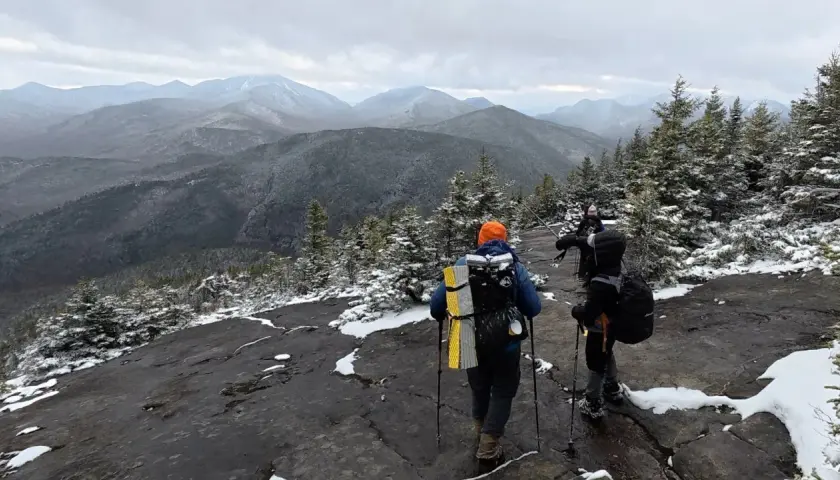 Hikers trek across a snowy mountain ridge, leaving no hiker trash behind. The vast, misty mountain range stretches into the distance under a cloudy sky.