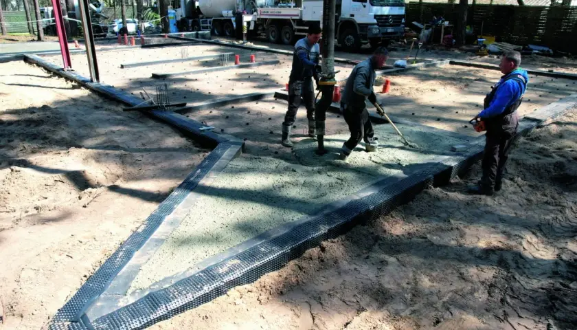Pracownicy wylewają beton na budowie, przygotowując podłoże pod posadzkę. Widać ciężarówkę z mieszanką betonową i sprzęt budowlany.