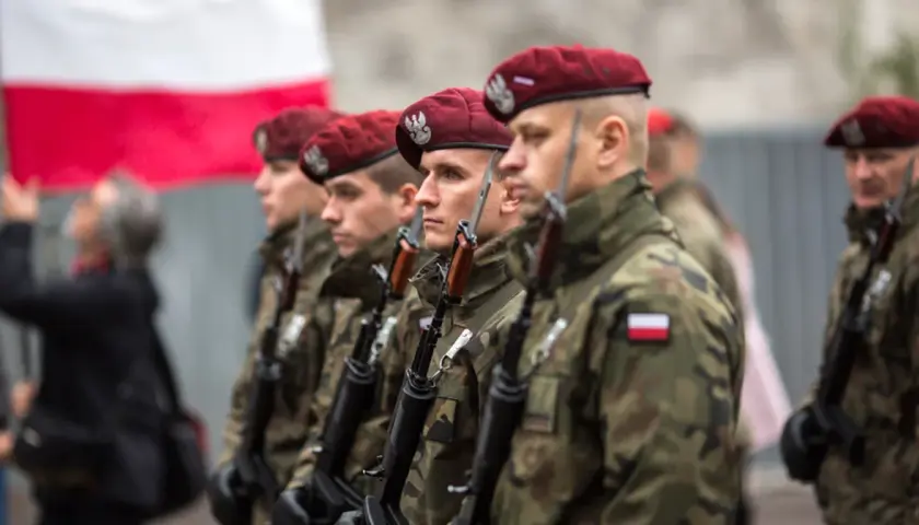 Żołnierze w bordowych beretach i mundurach moro, z karabinami, stoją w szeregu. W tle polska flaga. To dumne kategorie wojskowe.